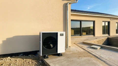 Unité extérieure de pompe à chaleur air/air installée contre un mur de façade enduite beige, posée sur silent-blocs, côté terrasse béton d'une maison neuve